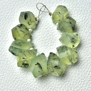 May include: A strand of faceted prehnite gemstone beads. The beads are a pale green colour with black inclusions, strung together with white beads and a silver-coloured wire. The gemstones have a polished, translucent appearance.