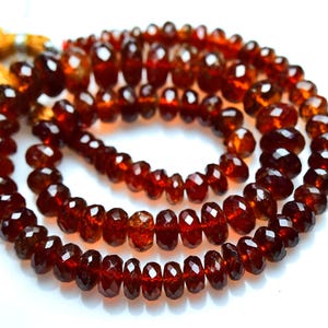 May include: A strand of deep red, faceted garnet beads arranged in a circular coil. The translucent, dark crimson gemstones feature a polished, multi-faceted surface that reflects light. A golden-brown silk tassel is attached to the end.