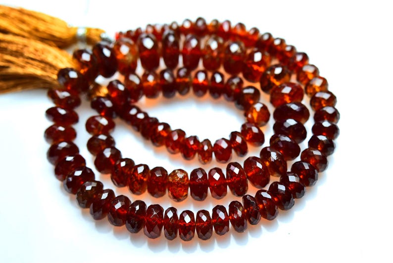 May include: A strand of deep red, faceted garnet beads arranged in a circular coil. The translucent, dark crimson gemstones feature a polished, multi-faceted surface that reflects light. A golden-brown silk tassel is attached to the end.