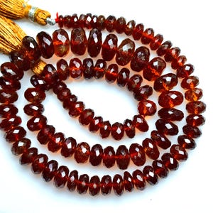May include: A strand of faceted, deep red garnet beads arranged in a circular coil. The beads are translucent with a polished finish, showing varied sizes and shapes. Two golden-yellow silk tassels are attached to the end of the bead strand.