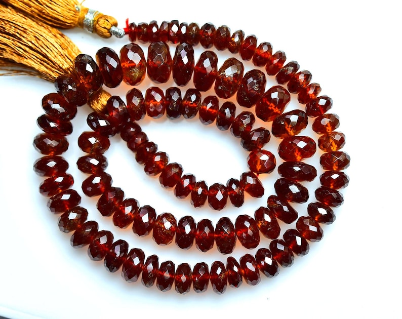 May include: A strand of faceted, deep red garnet beads arranged in a circular coil. The beads are translucent with a polished finish, showing varied sizes and shapes. Two golden-yellow silk tassels are attached to the end of the bead strand.