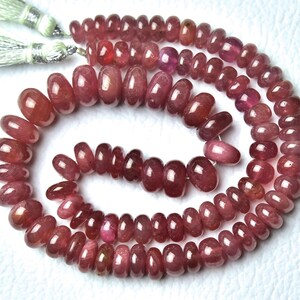 May include: A necklace or bracelet made of polished, oval-shaped, deep red gemstones. The beads vary in size and are strung together. The tassels at the top are a light green colour.