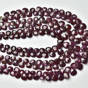 Pode incluir: Um fio de contas de rubi vermelho escuro, cada conta é facetada e tem o formato de um coração. As contas são enfiadas em um fio transparente e estão dispostas em um padrão solto e fluido.