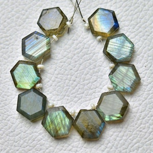 May include: A strand of iridescent labradorite gemstone beads. Each hexagonal bead is a mix of gray, green, and blue, strung together with white spacers. The beads are on a white surface.