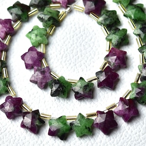 Könnte beinhalten: Eine Strähne aus sternförmigen grünen und violetten Edelsteinperlen, die durch goldene Abstandshalter getrennt sind. Die Perlen sind facettiert und haben ein sternförmiges Muster.