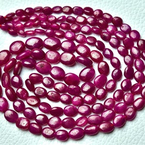 May include: A strand of ruby gemstone beads, each bead is a smooth, oval shape with a deep red colour. The beads are arranged in a loose coil on a white surface.