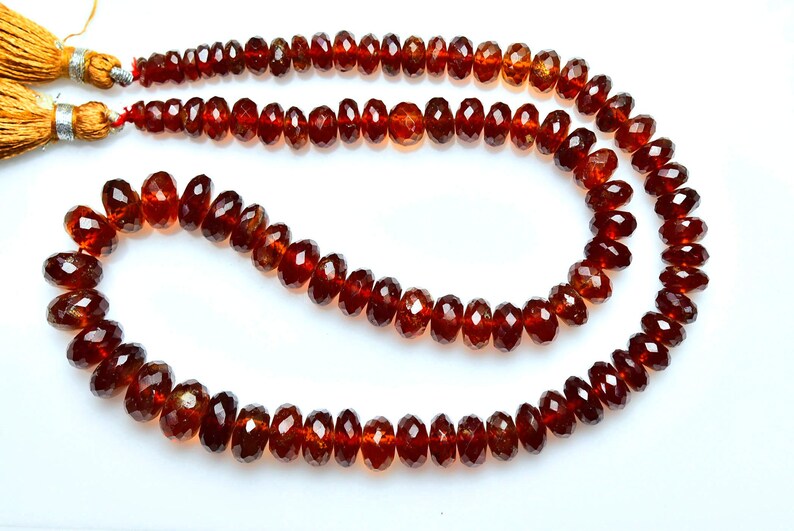 May include: A strand of faceted, deep red garnet beads arranged in a curved shape. The beads are translucent and have a polished finish. At one end of the strand, there are two golden-colored tassels attached with silver-toned metal caps.