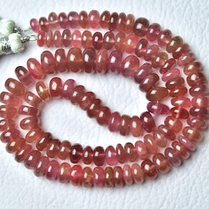 May include: A necklace of polished, oval-shaped beads in shades of pink and red. The beads are strung together, with a tassel at each end. The beads are approximately 0.6 cm in diameter. The necklace is displayed on a white surface.