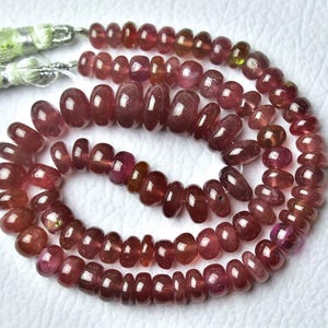May include: A necklace of polished, oval-shaped beads in shades of red and burgundy. The beads are strung together, with a tassel at the end. The beads are likely gemstones, and the overall aesthetic is elegant and natural.