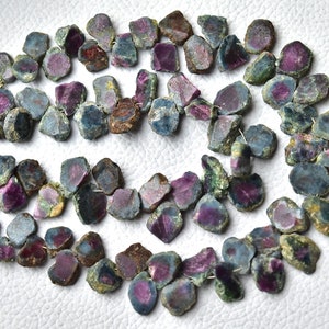 Pode incluir: Um fio de contas de rubi natural bruto com uma mistura de tons verdes, azuis e rosados. As contas são irregulares em forma e tamanho e têm um acabamento natural não polido.