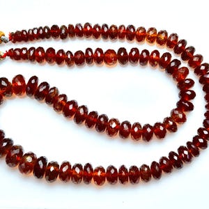 May include: A strand of deep red, faceted garnet beads arranged in a curved shape, featuring a polished finish and a translucent quality. Two golden-brown silk tassels are attached to the ends of the string, secured with silver-toned metal caps.