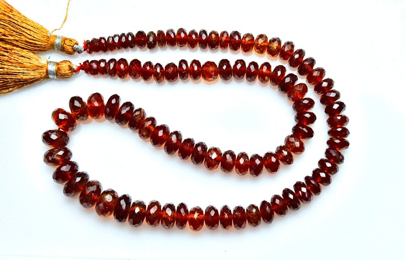 May include: A strand of deep red, faceted garnet beads arranged in a curved shape, featuring a polished finish and a translucent quality. Two golden-brown silk tassels are attached to the ends of the string, secured with silver-toned metal caps.