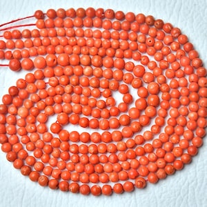 Op de afbeelding: Een verzameling kleine, ronde, oranje koraalkralen geregen op rood draad. De kralen zijn gerangschikt in een spiraalpatroon, met verschillende maten. Het koraal heeft een natuurlijk, getextureerd uiterlijk en de presentatie is netjes.