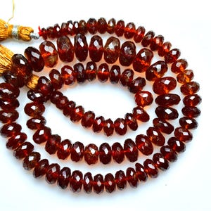 May include: A strand of deep red, faceted garnet beads arranged in a circular coil. The beads are translucent and have a polished finish. Two golden-brown silk tassels are attached to the end of the string, providing a contrast to the dark red stones.