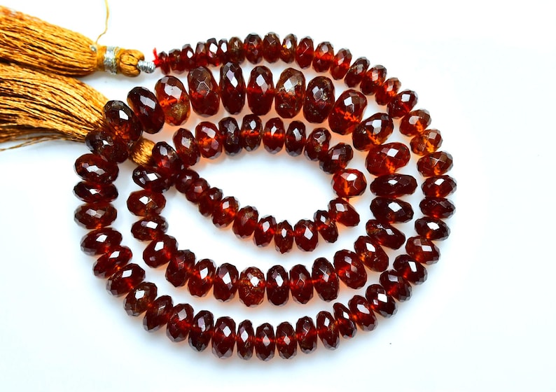 May include: A strand of deep red, faceted garnet beads arranged in a circular coil. The beads are translucent and have a polished finish. Two golden-brown silk tassels are attached to the end of the string, providing a contrast to the dark red stones.