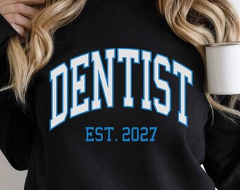 May include: A black long-sleeve sweatshirt featuring the word DENTIST in a large, arched, light blue font with white outlines. Below the word, the text EST. 2027 is printed in a matching light blue font. The garment is worn with a white mug.
