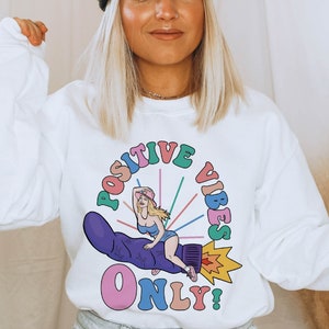 May include: White sweatshirt with a colorful graphic design featuring a woman riding a purple rocket with the text "POSITIVE VIBES ONLY!"