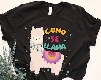 Puede incluir: Camiseta negra con una llama de dibujos animados que lleva una silla de montar con estampado colorido y el texto "COMO SE LLAMA" en español.