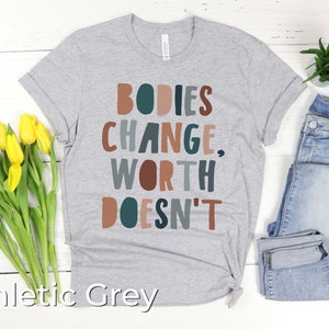 May include: A heather grey t-shirt with the text "BODIES CHANGE, WORTH DOESN'T" in colorful letters. The shirt is laid flat on a white wooden surface with a bouquet of yellow tulips and a pair of blue jeans.