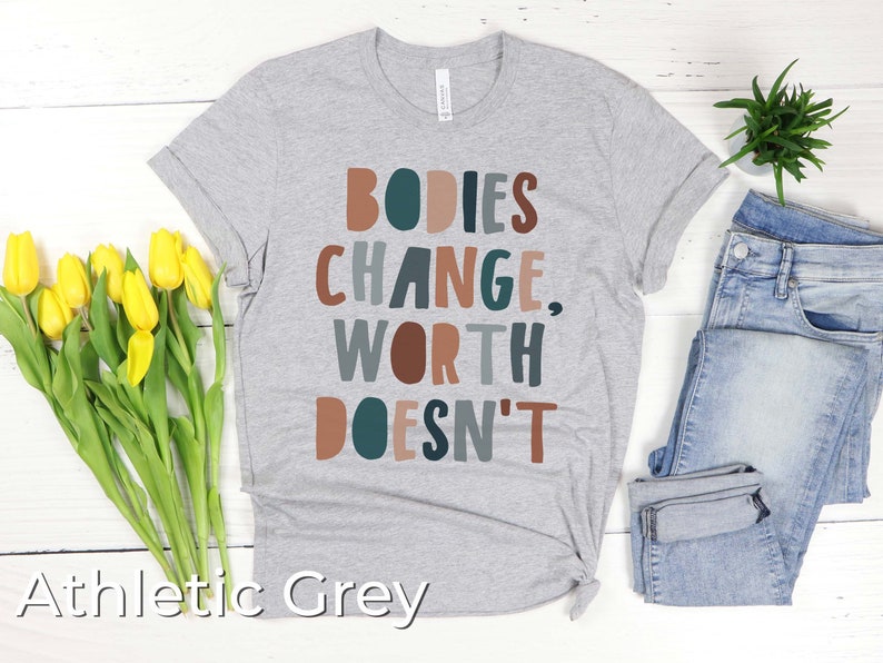 May include: A heather grey t-shirt with the text "BODIES CHANGE, WORTH DOESN'T" in colorful letters. The shirt is laid flat on a white wooden surface with a bouquet of yellow tulips and a pair of blue jeans.