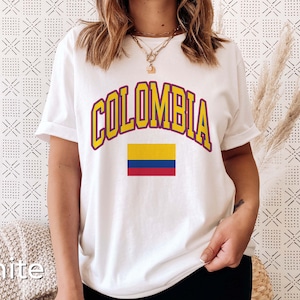 K&ouml;nnte beinhalten: Wei&szlig;es T-Shirt mit dem Wort "COLOMBIA" in gelben Buchstaben mit rotem Rand und einer kleinen kolumbianischen Flagge darunter.