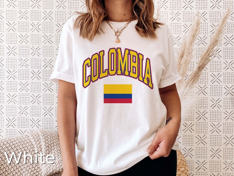 K&ouml;nnte beinhalten: Wei&szlig;es T-Shirt mit dem Wort "COLOMBIA" in gelben Buchstaben mit rotem Rand und einer kleinen kolumbianischen Flagge darunter.