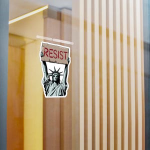 Puede incluir: Una pegatina con una ilustraci&oacute;n de dibujos animados de la Estatua de la Libertad sosteniendo un cartel que dice "RESIST" en letras rojas.