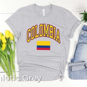 K&ouml;nnte beinhalten: Ein graues T-Shirt mit dem Wort "COLOMBIA" in gelben Buchstaben mit rotem Rand. Die kolumbianische Flagge ist unter dem Wort gedruckt. Das T-Shirt liegt auf einem wei&szlig;en Holzgrund.