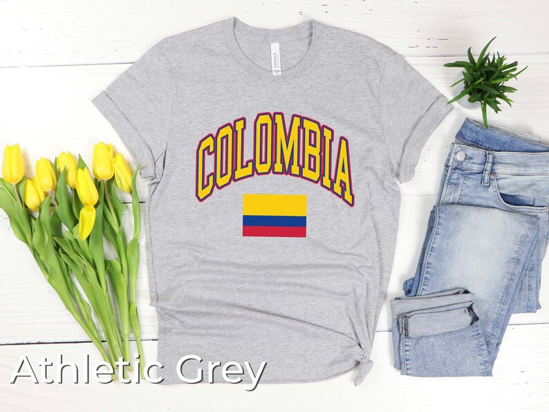 K&ouml;nnte beinhalten: Ein graues T-Shirt mit dem Wort "COLOMBIA" in gelben Buchstaben mit rotem Rand. Die kolumbianische Flagge ist unter dem Wort gedruckt. Das T-Shirt liegt auf einem wei&szlig;en Holzgrund.