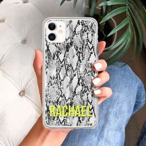 Puede incluir: Una funda transparente para teléfono con un patrón de piel de serpiente en blanco y negro. La funda tiene el nombre "RACHAEL" impreso en letras amarillas en la parte inferior.