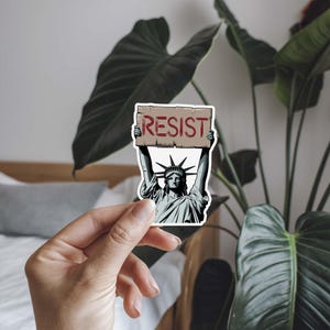 Puede incluir: Una pegatina con una ilustraci&oacute;n de dibujos animados de la Estatua de la Libertad sosteniendo un cartel que dice "RESIST".