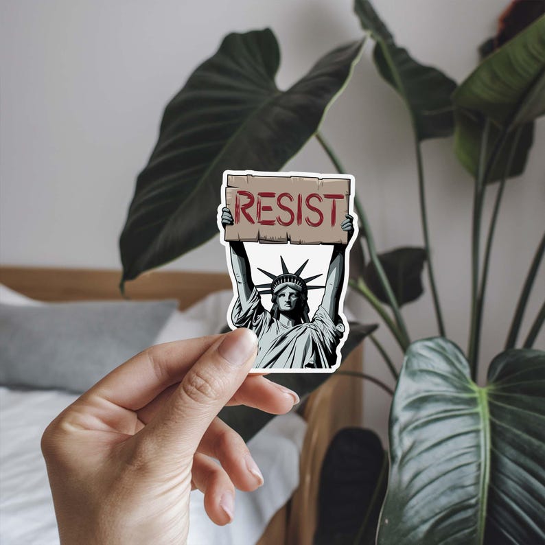 Puede incluir: Una pegatina con una ilustraci&oacute;n de dibujos animados de la Estatua de la Libertad sosteniendo un cartel que dice "RESIST".