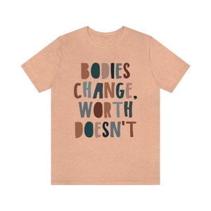 May include: A peach-colored t-shirt with the text "BODIES CHANGE, WORTH DOESN'T" in colorful, stylized lettering.