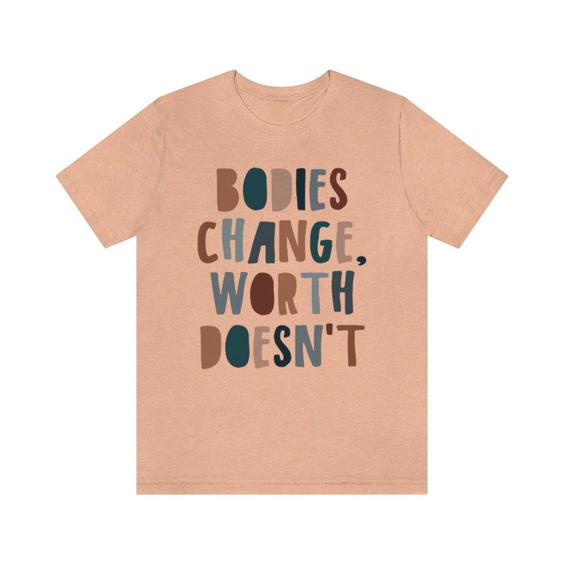 May include: A peach-colored t-shirt with the text "BODIES CHANGE, WORTH DOESN'T" in colorful, stylized lettering.