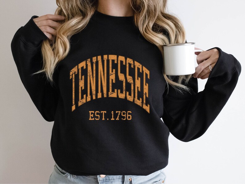 Puede incluir: Sudadera negra con un gr&aacute;fico naranja desgastado que dice "TENNESSEE EST. 1796".