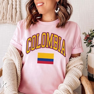 K&ouml;nnte beinhalten: Rosafarbenes T-Shirt mit dem Wort "COLOMBIA" in gelben Buchstaben mit blauem Umriss und einer kleinen Grafik der kolumbianischen Flagge.