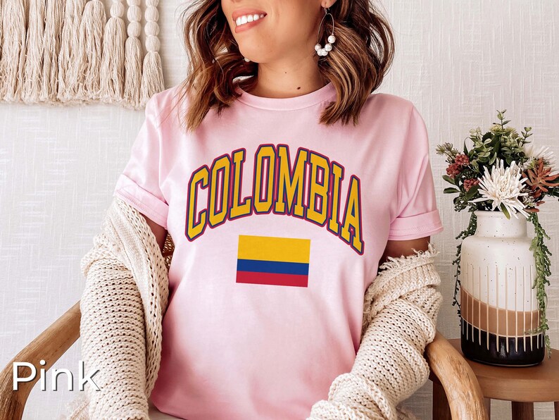 K&ouml;nnte beinhalten: Rosafarbenes T-Shirt mit dem Wort "COLOMBIA" in gelben Buchstaben mit blauem Umriss und einer kleinen Grafik der kolumbianischen Flagge.