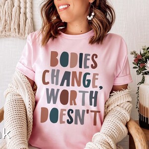 May include: Pink t-shirt with the text "BODIES CHANGE, WORTH DOESN'T" in colorful, stylized lettering.