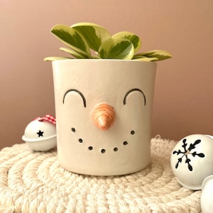 Puede incluir: Maceta de cerámica blanca con forma de cara de muñeco de nieve con una boca sonriente, ojos cerrados y una nariz de zanahoria naranja. La maceta está llena de una planta verde y blanca variegada.