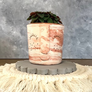 Könnte beinhalten: Dekorativer Keramik-Pflanztopf mit Gesichtsdesign, mit einem rosa-weißen Marmormuster. Der Pflanztopf enthält eine kleine grüne Pflanze. Er steht auf einem grauen, gewellten Untersetzer, auf einem cremefarbenen Makramee-Platzset.