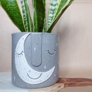 Könnte beinhalten: Ein grauer Betontopf mit einem weißen Halbmond-Design. Der Mond hat ein lächelndes Gesicht mit geschlossenen Augen und eine strukturierte Oberfläche. Der Topf hat eine gesprenkelte graue Oberfläche und weiße Sterne.