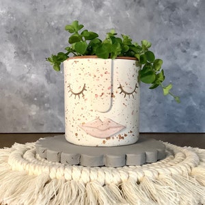 Puede incluir: Maceta de cerámica blanca con un diseño de cara rosa y dorada. La maceta tiene los ojos cerrados, una nariz y una boca rosa. Una planta suculenta verde está creciendo en la maceta. La maceta está sentada sobre un posavasos circular gris y blanco.