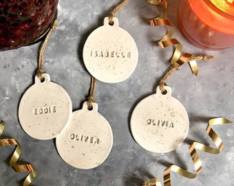 Könnte beinhalten: Vier weiße Keramik-Ornamente mit goldener Beschriftung. Die Ornamente sind in Form von Christbaumkugeln gestaltet und tragen die Namen "Isabelle", "Eddie", "Oliver" und "Olivia".