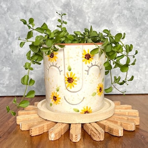 Può includere: Un vaso di ceramica bianca con un viso sorridente dipinto su di esso. Il vaso è decorato con girasoli gialli e foglie verdi. Una piccola pianta verde sta crescendo nel vaso. Il vaso è appoggiato su un sottopentola di legno a forma di sole.