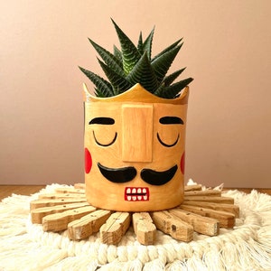 May include: A hand-painted ceramic planter shaped like a face with a gold crown, closed eyes, a black mustache, and red cheeks. A green succulent plant is growing in the planter.