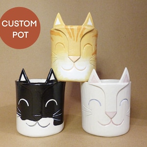 May include: Three ceramic cat planter pots, one orange, one black and white, and one white with pink accents. Each pot has a smiling cat face with ears.
