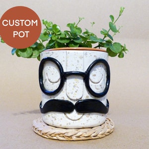 Könnte beinhalten: Ein weißer Keramiktopf mit braunem Rand, der mit einem schwarzen Schnurrbart, einer Brille und einem lächelnden Gesicht verziert ist. Der Topf ist mit einer grünen Pflanze gefüllt und steht auf einem geflochtenen Untersetzer.