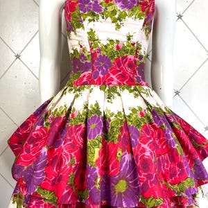 K&ouml;nnte beinhalten: Ein tr&auml;gerloses Kleid mit wei&szlig;em Hintergrund und einem Blumendruck in Rosa-, Lila- und Gr&uuml;nt&ouml;nen. Das Kleid hat ein tailliertes Oberteil und einen vollen Rock mit mehreren R&uuml;schenlagen.