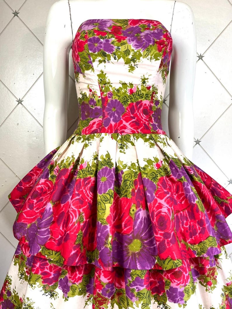 K&ouml;nnte beinhalten: Ein tr&auml;gerloses Kleid mit wei&szlig;em Hintergrund und einem Blumendruck in Rosa-, Lila- und Gr&uuml;nt&ouml;nen. Das Kleid hat ein tailliertes Oberteil und einen vollen Rock mit mehreren R&uuml;schenlagen.