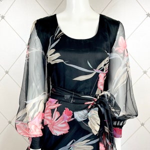 Peut inclure: Robe noire &agrave; imprim&eacute; floral avec manches transparentes et ceinture assortie. La robe pr&eacute;sente une encolure arrondie et une jupe fluide. Le motif floral comprend des accents roses, blancs et gris. La robe est pr&eacute;sent&eacute;e sur un mannequin.